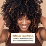 Person with voluminous dark curly hair with bronze highlights, hands in hair, smiling against beige background.