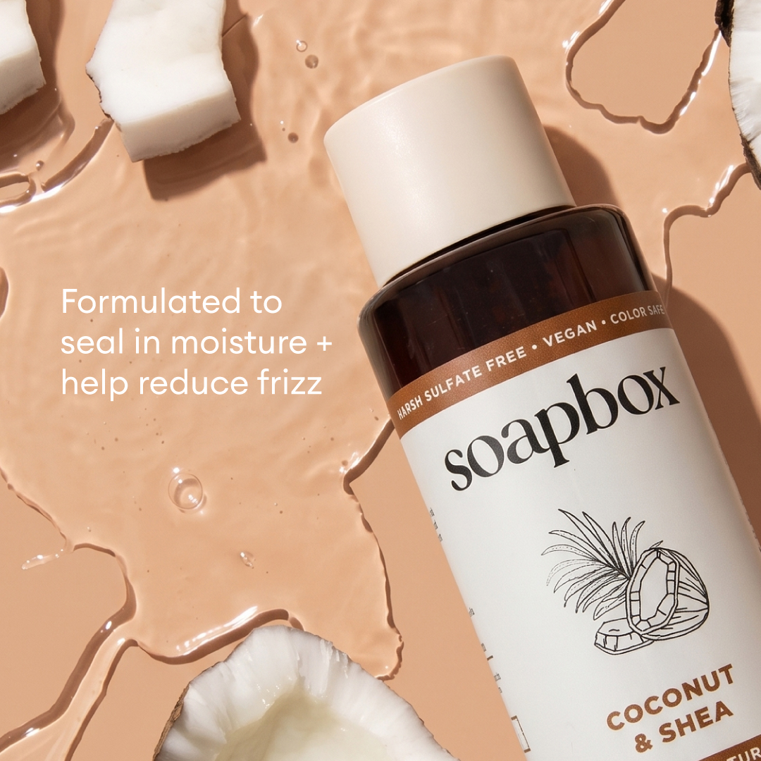 Soapbox Coconut & Shea shampoo bottle with white cap surrounded by coconut pieces and beige liquid on tan background.
