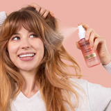 Person with reddish-brown hair holding a pink spray bottle labeled "Lychee Hibiscus" against a pink background.