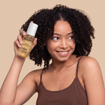 Spray bottle with white and gold label held next to curly dark hair against beige background.