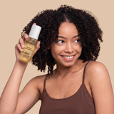 Spray bottle with white and gold label held next to curly dark hair against beige background.
