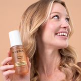 Woman holding a bronze-toned spray bottle labeled Vetiver Amber Hair Fragrance Finishing Mist with wavy blonde hair.