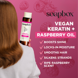 Person with brown wavy hair holding a Soapbox shampoo bottle with keratin and raspberry oil formula against pink background.