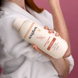 Person holding cream-colored body wash bottle with vanilla and pro-bio label against pink background.