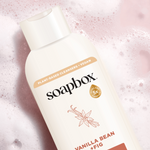 Soapbox body wash bottle with cream-colored label and vanilla bean plus fig text on pink sparkly background.
