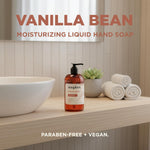 Amber bottle of Soapbox Vanilla Bean hand soap with pump dispenser on bathroom counter next to white bowl, towels, and plant.