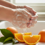 Hands lathered with soap and water, with orange slices and whole oranges displayed below.