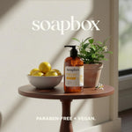 Soapbox hand soap bottle with black pump on wooden table beside bowl of yellow lemons and potted herbs.
