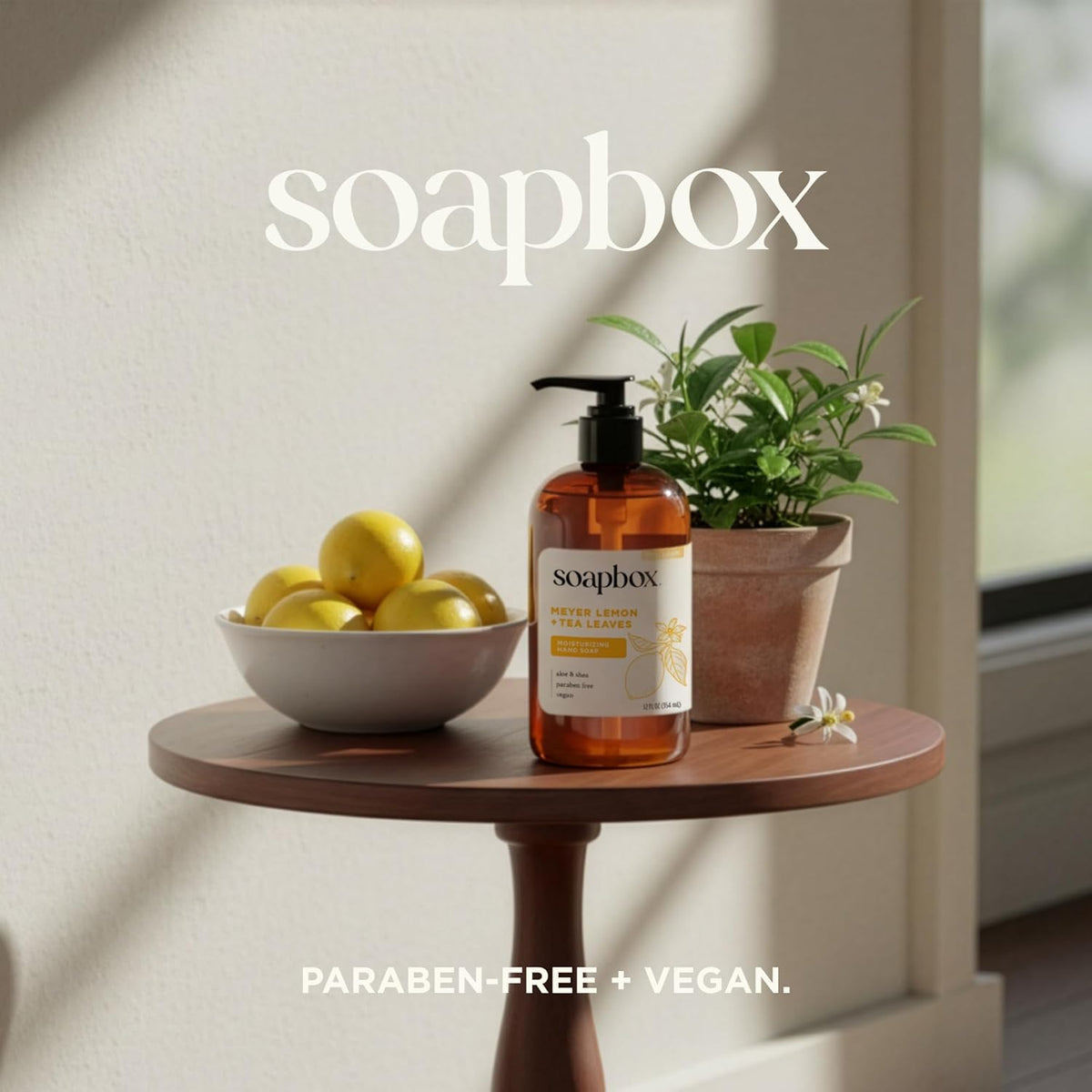 Soapbox hand soap bottle with black pump on wooden table beside bowl of yellow lemons and potted herbs.
