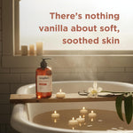 Amber liquid hand soap bottle with black pump dispenser on wooden tub ledge beside candles and vanilla flower.