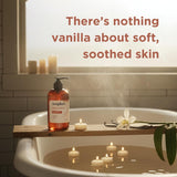 Amber liquid hand soap bottle with black pump dispenser on wooden tub ledge beside candles and vanilla flower.