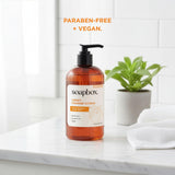 Soapbox sweet orange citrus liquid hand soap in amber bottle with black pump, displayed with white towels and potted plant.