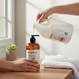Hand pouring Soapbox coconut milk and sandalwood liquid hand soap from gallon refill jug into amber glass dispenser bottle.