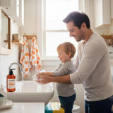Man and toddler washing hands at sink with orange-labeled liquid hand soap dispenser nearby.