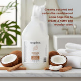 Soapbox gallon refill bottle surrounded by halved coconuts, cinnamon sticks, and folded white towels on marble surface.