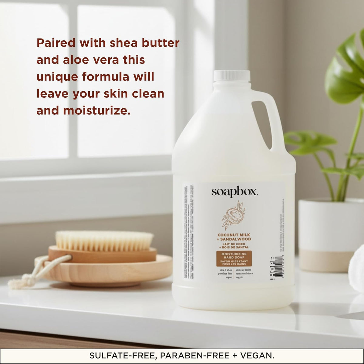 Large white refillable hand soap bottle labeled "Coconut Milk + Sandalwood" displayed with natural bristle brush and potted p