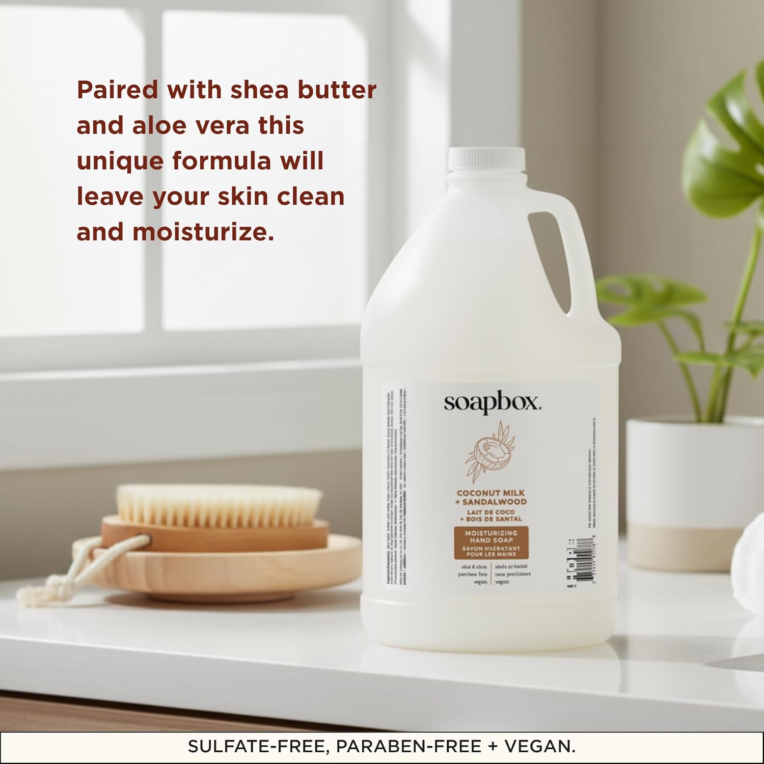 Large white refillable hand soap bottle labeled "Coconut Milk + Sandalwood" displayed with natural bristle brush and potted p