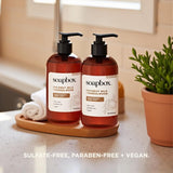 Two amber Soapbox hand soap pump bottles with black pumps on wooden tray next to potted plant and rolled towel.
