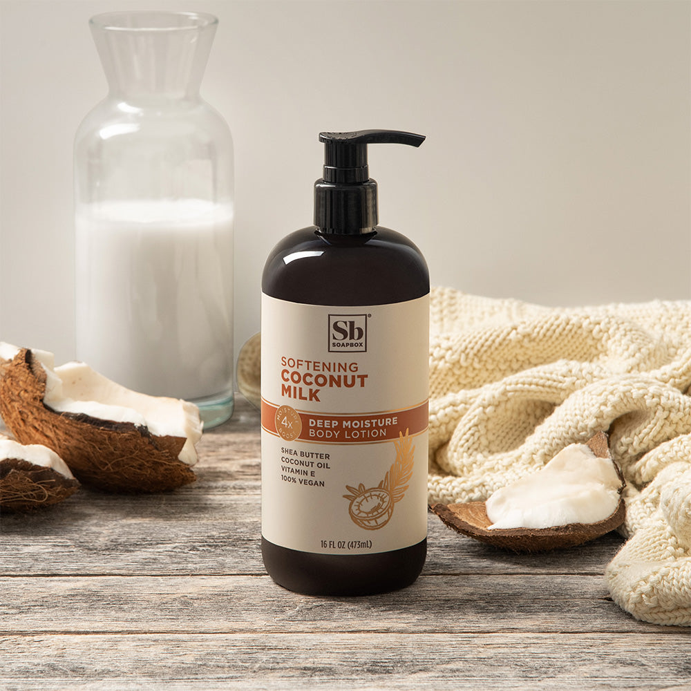 Black pump bottle of coconut milk body lotion surrounded by coconut halves, cream fabric, and clear glass bottle on wooden su