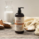 Black pump bottle of coconut milk body lotion surrounded by coconut halves, cream fabric, and clear glass bottle on wooden su