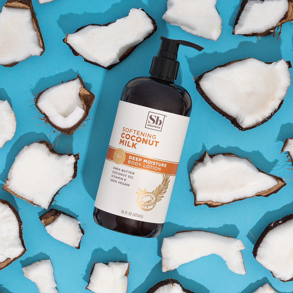 Black pump bottle of coconut milk body lotion surrounded by halved coconuts on bright blue background.