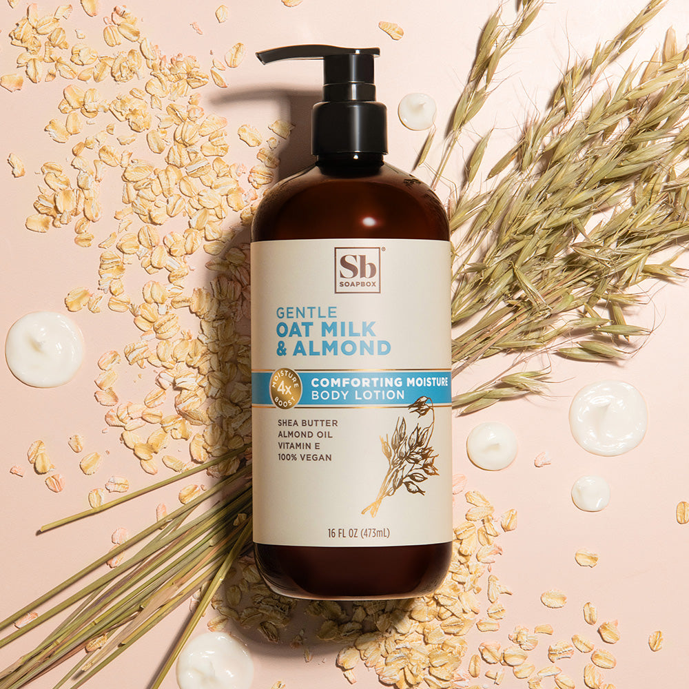 Brown pump bottle of body lotion surrounded by oat flakes, almond pieces, dried wheat stalks, and white cream dollops on pink