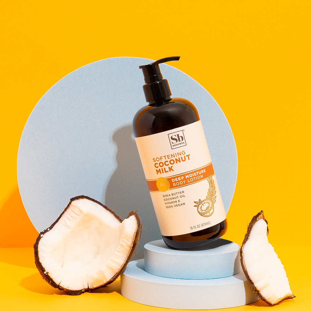 Dark brown bottle with pump dispenser of Softening Coconut Milk Deep Moisture Body Lotion, flanked by fresh coconut halves on