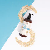 Amber glass pump bottle of oat milk and almond body lotion surrounded by scattered oats on blue and white background.
