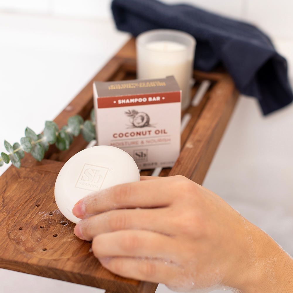 Hand holding white coconut oil shampoo bar on wooden tray with product packaging, candle, and greenery.