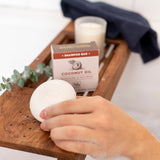 Hand holding white coconut oil shampoo bar on wooden tray with product packaging, candle, and greenery.