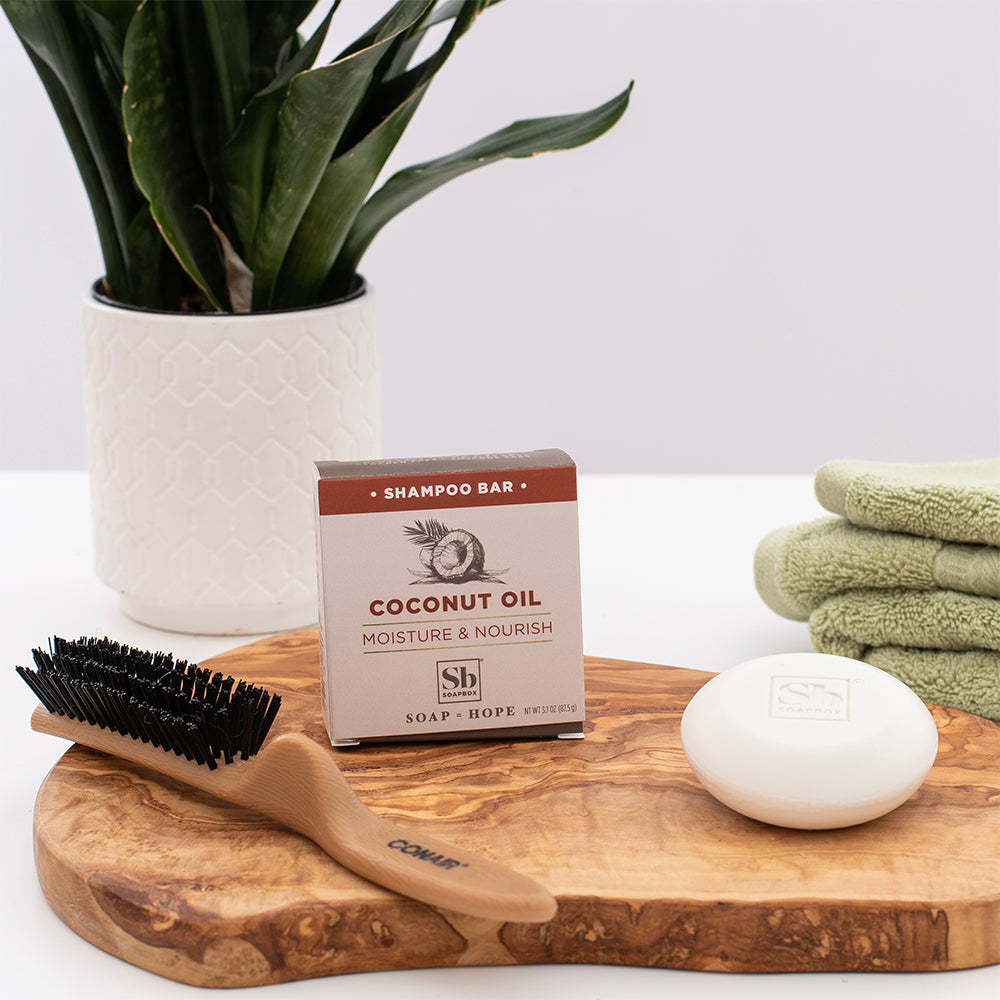 Coconut Oil Moisture & Nourish shampoo bar displayed on wooden tray with brush, soap dish, folded towels, and potted plant.