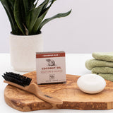 Coconut Oil Moisture & Nourish shampoo bar displayed on wooden tray with brush, soap dish, folded towels, and potted plant.