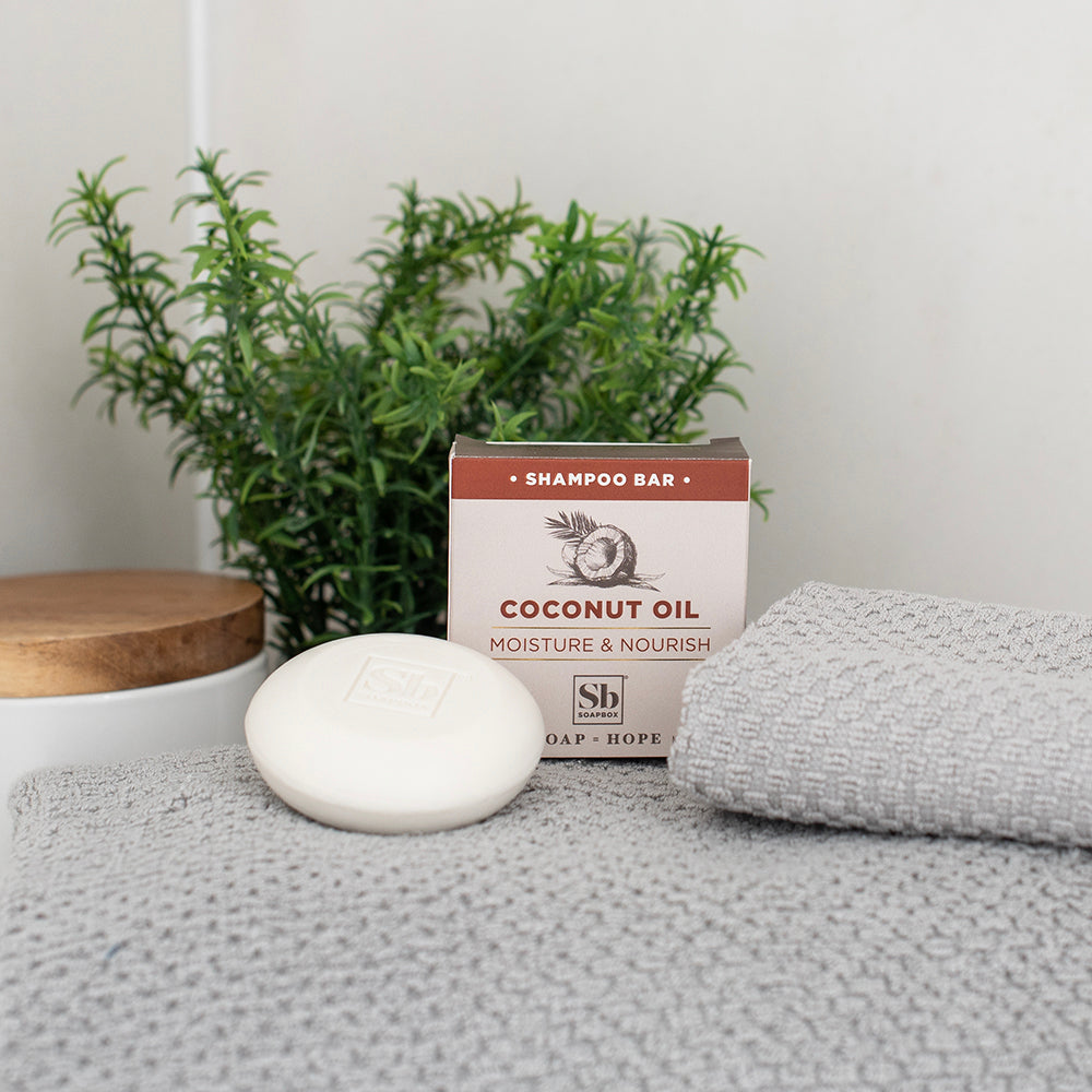 Cream-colored shampoo bar with coconut oil packaging, displayed with folded gray towel and green plant.