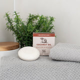 Cream-colored shampoo bar with coconut oil packaging, displayed with folded gray towel and green plant.