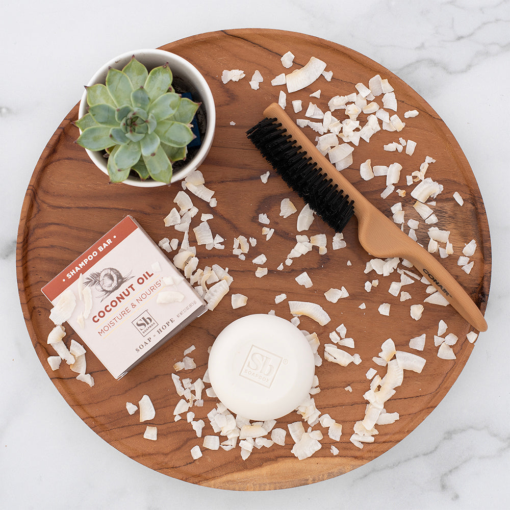 Coconut oil shampoo bar with packaging, wooden brush, shredded coconut, and succulent plant on wooden tray.
