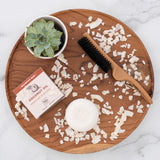 Coconut oil shampoo bar with packaging, wooden brush, shredded coconut, and succulent plant on wooden tray.