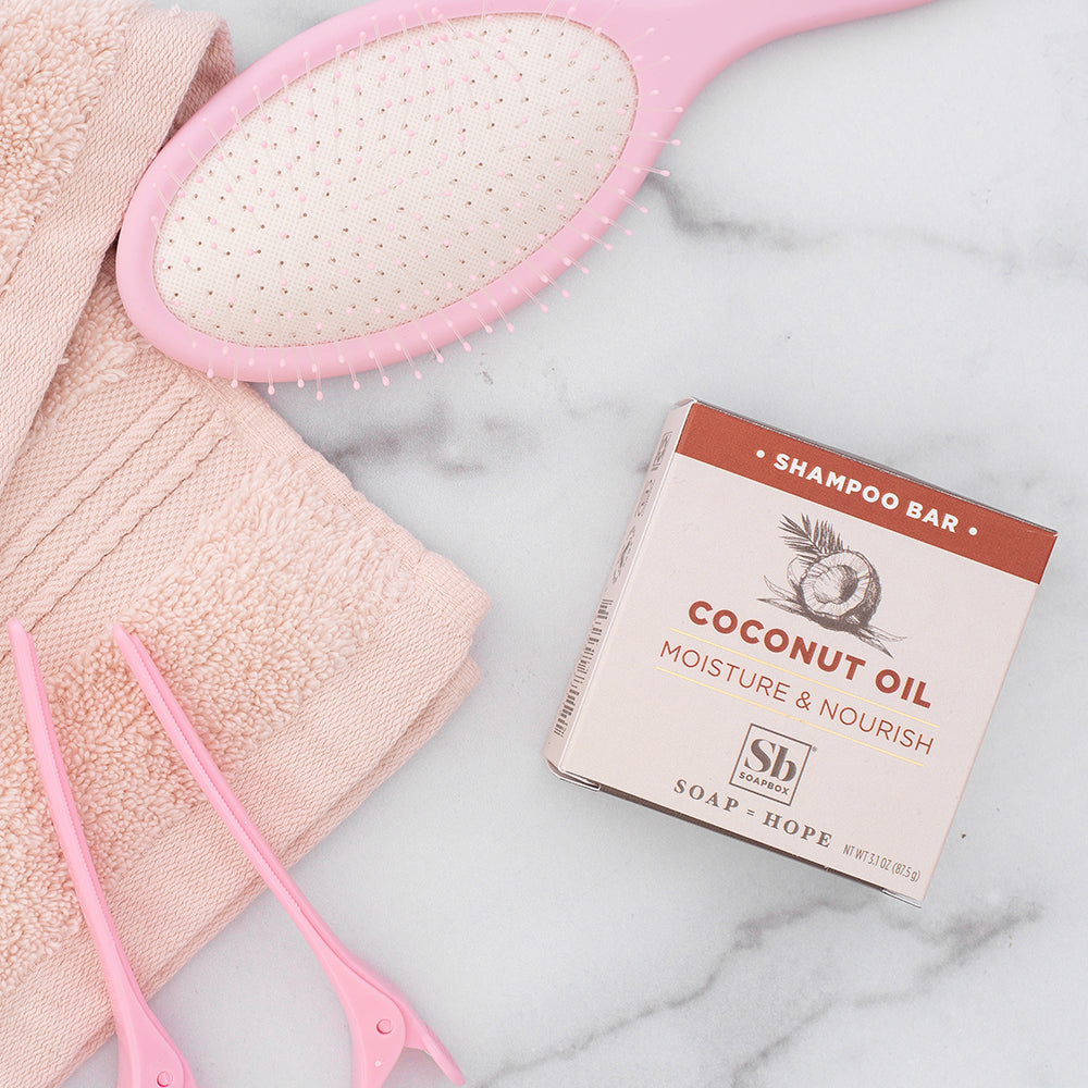 Coconut Oil Moisture & Nourish shampoo bar displayed next to pink brush, towel, and scissors on marble surface.