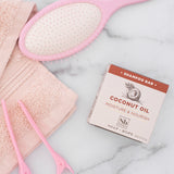 Coconut Oil Moisture & Nourish shampoo bar displayed next to pink brush, towel, and scissors on marble surface.