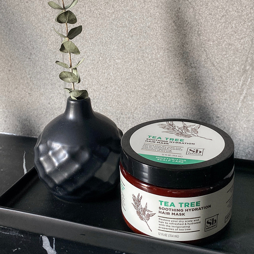 Tea Tree Soothing Hydration Hair Mask in black jar with white label, displayed next to black vase with green leaves.