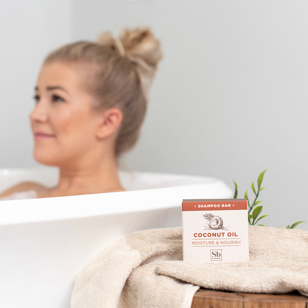 Coconut Oil Moisture & Nourish shampoo bar on beige towel with green plant sprig, woman in bathtub background.